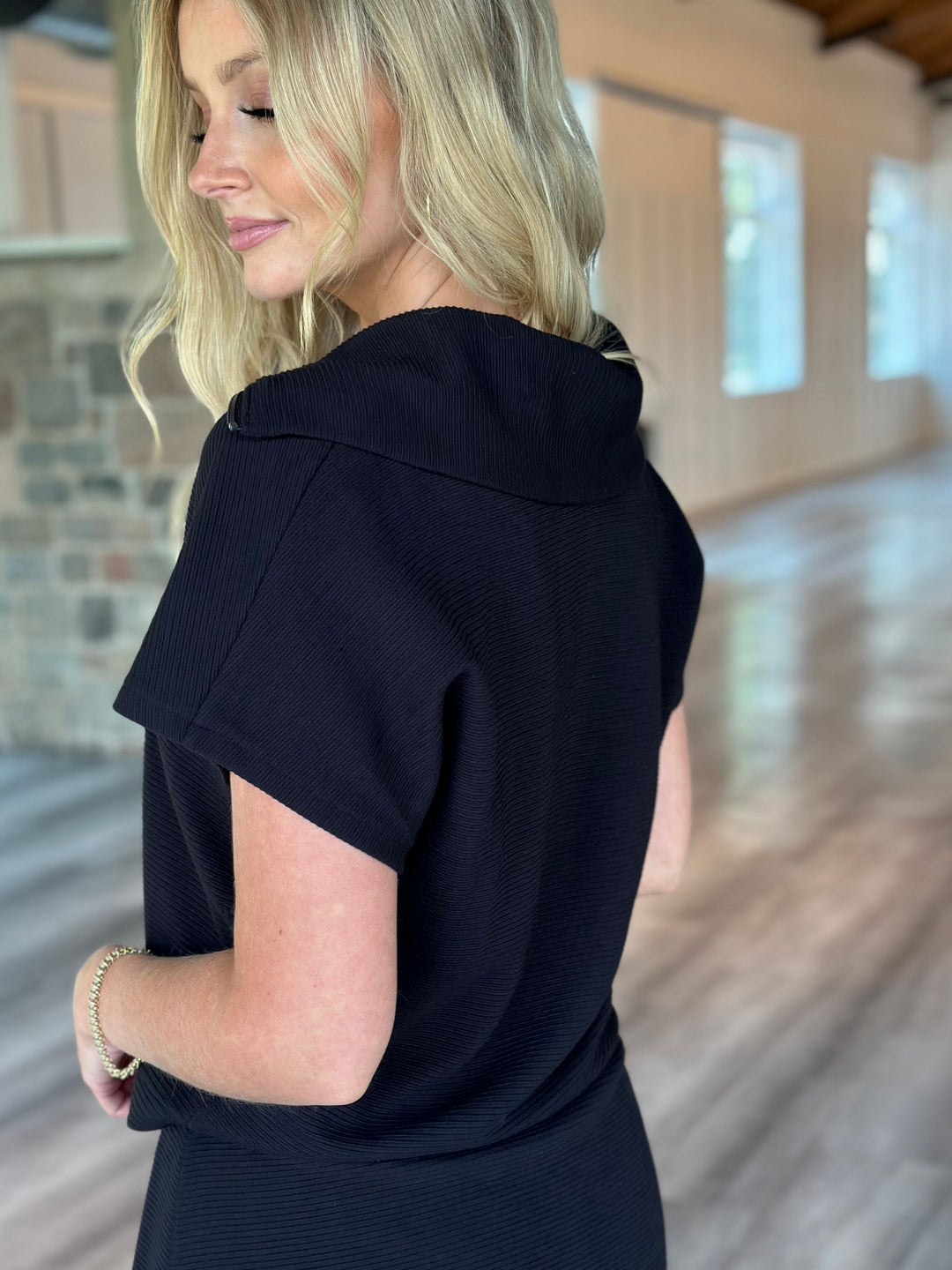 A woman wearing a black short-sleeve top with a wide collar, shown from a side-back angle in a bright indoor space with wood floors and windows.