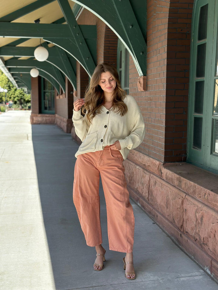 A woman stands on a covered outdoor walkway wearing a cream button-front cardigan and wide-leg cropped terracotta pants, with her hands in the pockets of her pants.