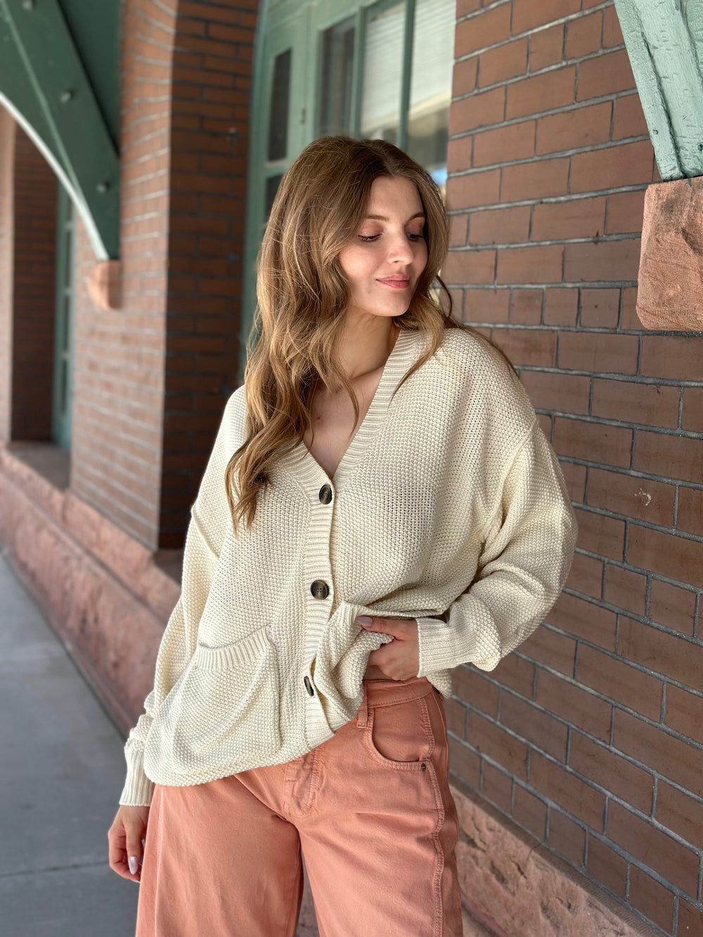 A woman wearing a cream waffle-knit button-up cardigan and peach pants, standing outdoors against a brick building wall.