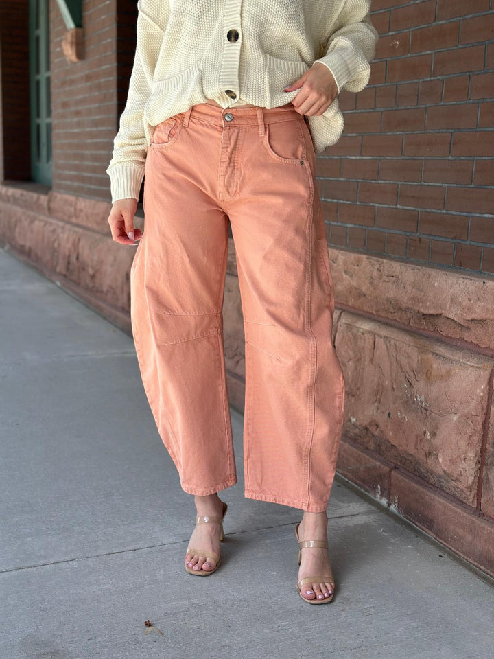 A person wearing wide-leg cropped peach-colored denim pants, a cream ribbed cardigan, and clear high-heeled sandals, standing outdoors against a brick wall.