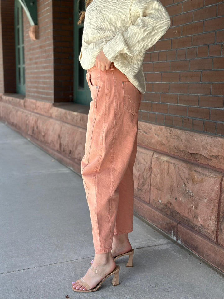 A person standing outdoors on a sidewalk beside a brick building, wearing a cream knit sweater, peach cropped pants, and clear heeled sandals.