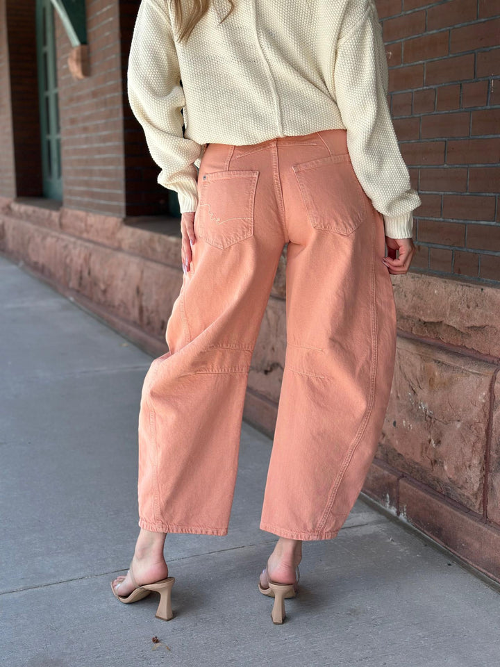 A person shown from the back wearing wide-leg cropped pants in a muted peach-pink color, paired with a cream knit sweater and beige heeled sandals, standing on a sidewalk next to a brick building.