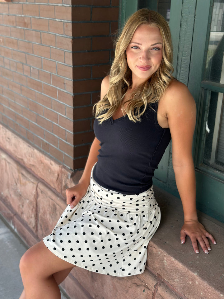 A woman with blonde hair sits on a stone ledge wearing a black tank top and a white A-line skirt with black polka dots, set against a brick and green door background.
