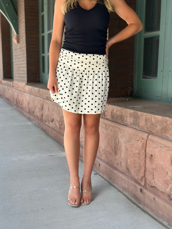 A woman wearing a black sleeveless top and a white mini skirt with black polka dots, standing outdoors on a sidewalk next to a brick building.
