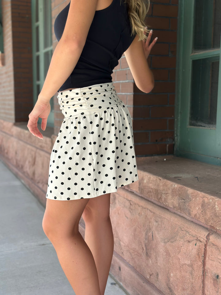 A woman wearing a black sleeveless top and a white high-waisted skater mini skirt with black polka dots, standing outdoors next to a brick building.