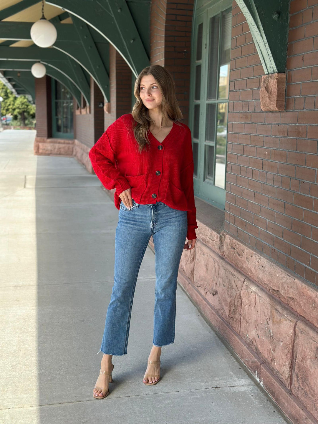 A woman stands on a sidewalk next to a brick building with green trim, wearing a bright red button-front cardigan, blue jeans, and clear high-heel sandals.
