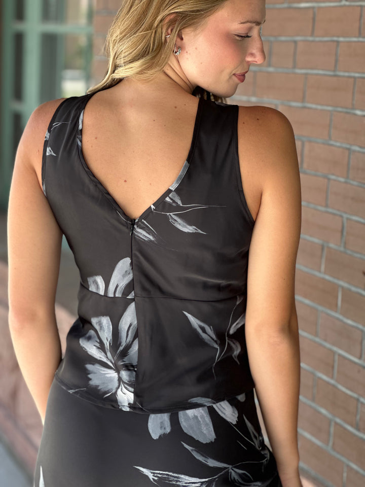 A woman wearing a sleeveless black dress with a gray and white floral print, shown from the back against a brick wall background.
