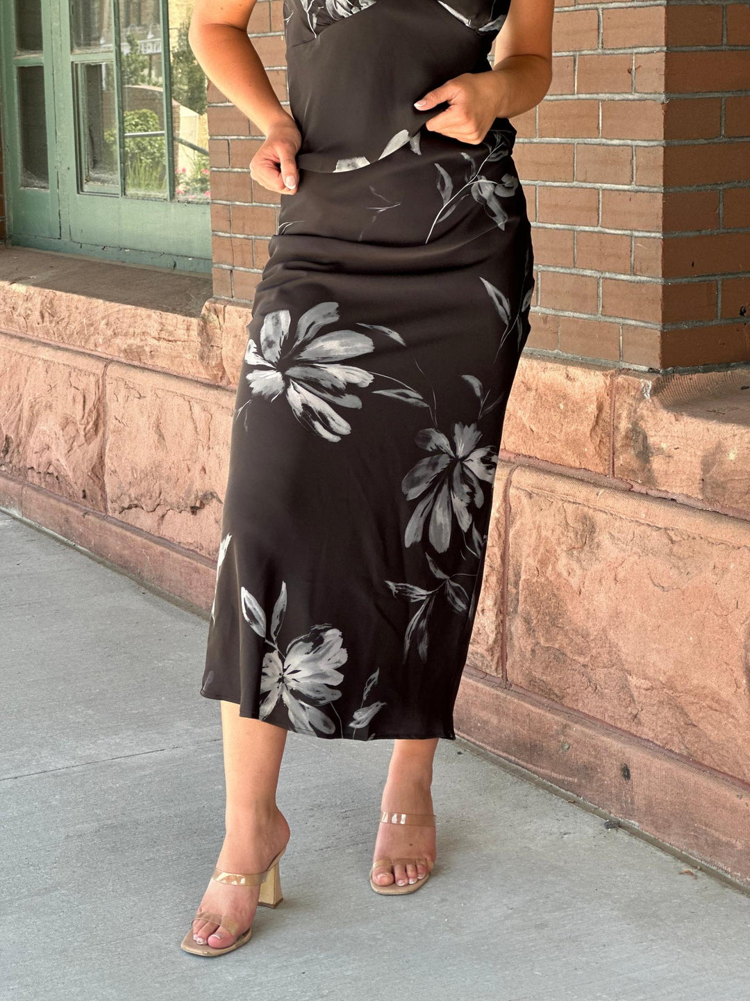 A woman wearing a black midi dress with a gray and white floral print, paired with clear strap heels, standing outdoors against a brick wall.