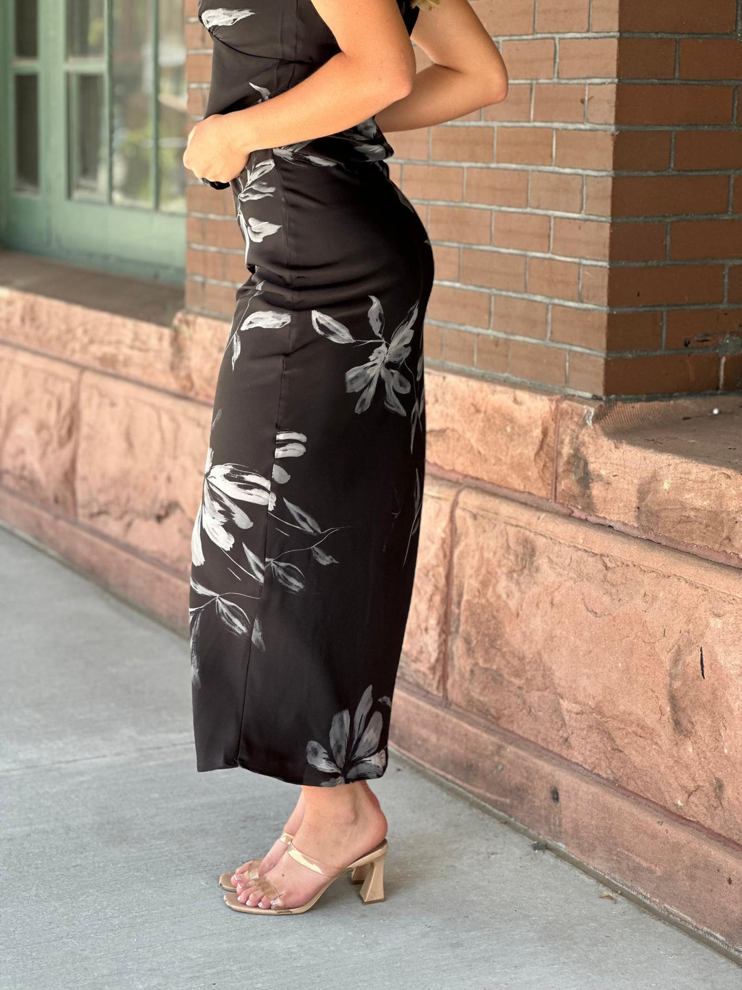 A woman wearing a black maxi skirt with a gray floral print, paired with clear heeled sandals, standing outdoors next to a brick wall.