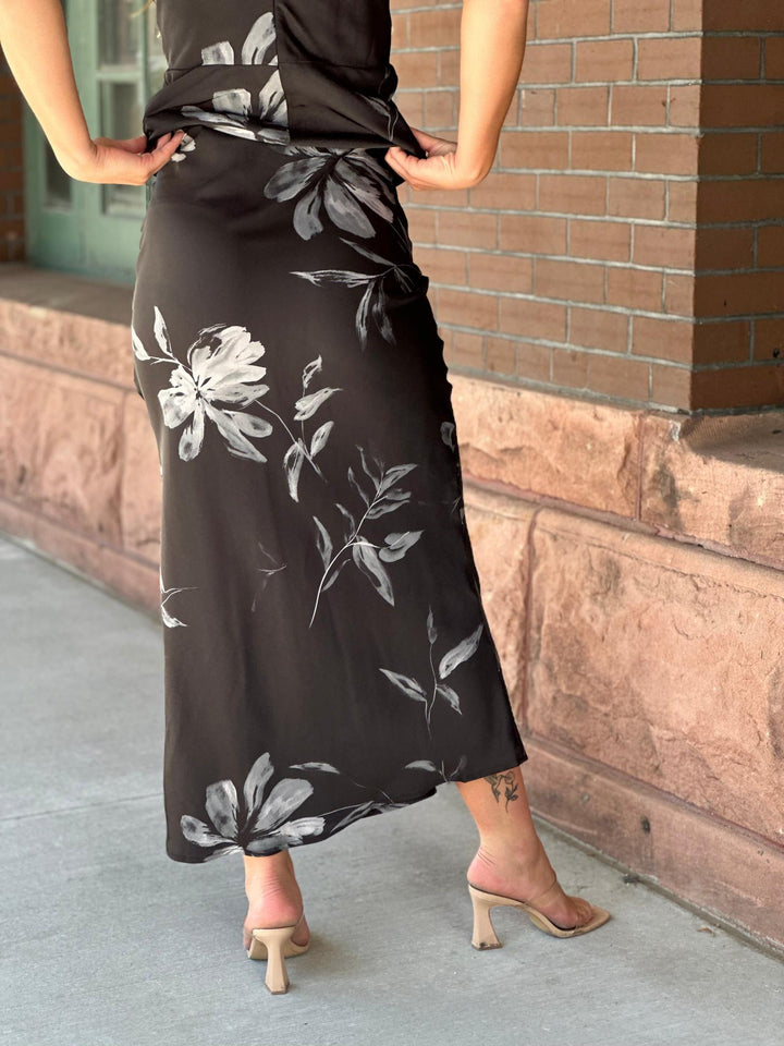 A woman wearing a black midi wrap skirt with a gray and white floral print, paired with beige heeled sandals, standing outdoors against a brick wall.