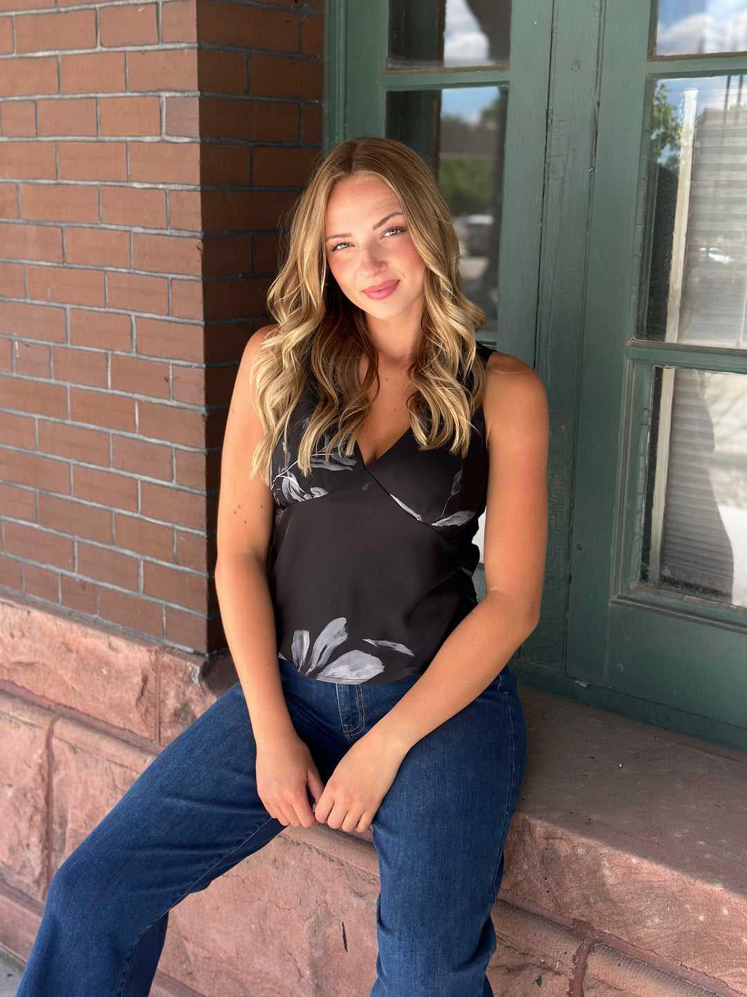 A woman with blonde wavy hair sits on a stone ledge wearing a black sleeveless tank top with a gray leaf pattern and blue jeans, positioned in front of a brick wall and a green wooden door.