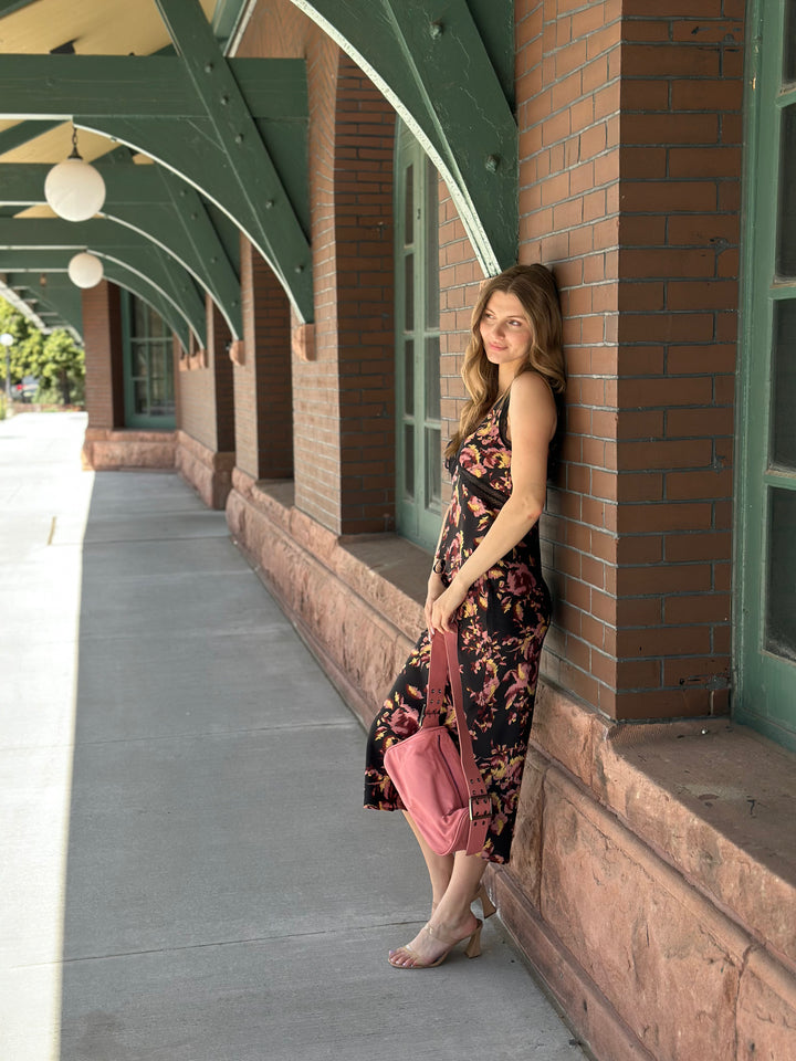 A woman stands barefoot against a brick building wall on a covered walkway, wearing a sleeveless black midi dress with a pink floral print and holding a pink handbag.