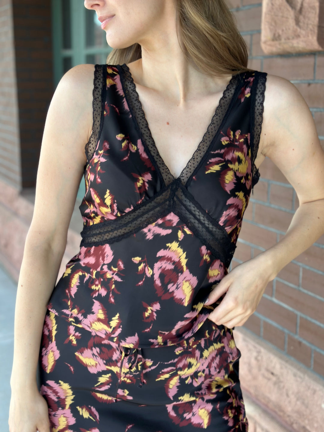 A woman wearing a sleeveless black satin dress with a pink and yellow floral print and black lace trim, standing against a brick wall.