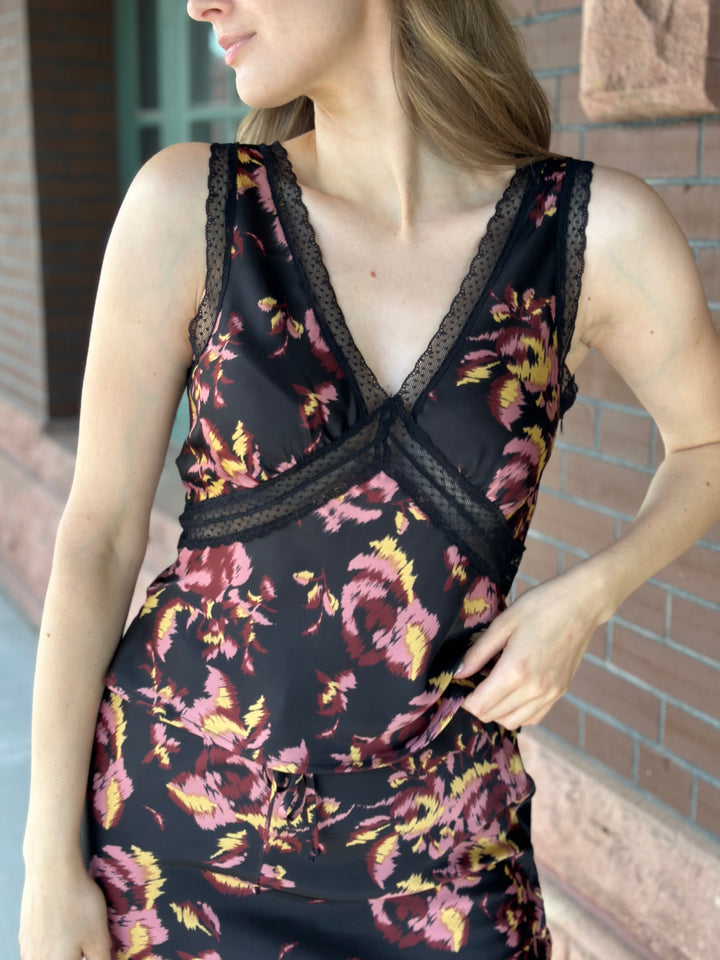 A woman wearing a sleeveless black satin dress with a pink and yellow floral print and black lace trim, standing against a brick wall.