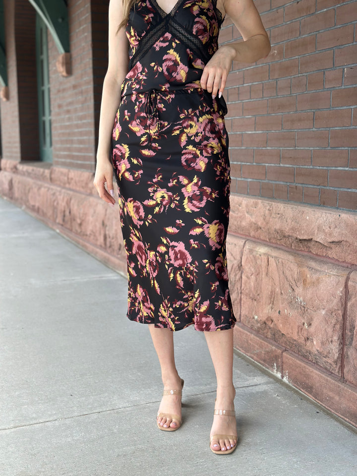 A woman wearing a sleeveless black midi dress with a pink and yellow floral print, standing on a sidewalk next to a brick building.
