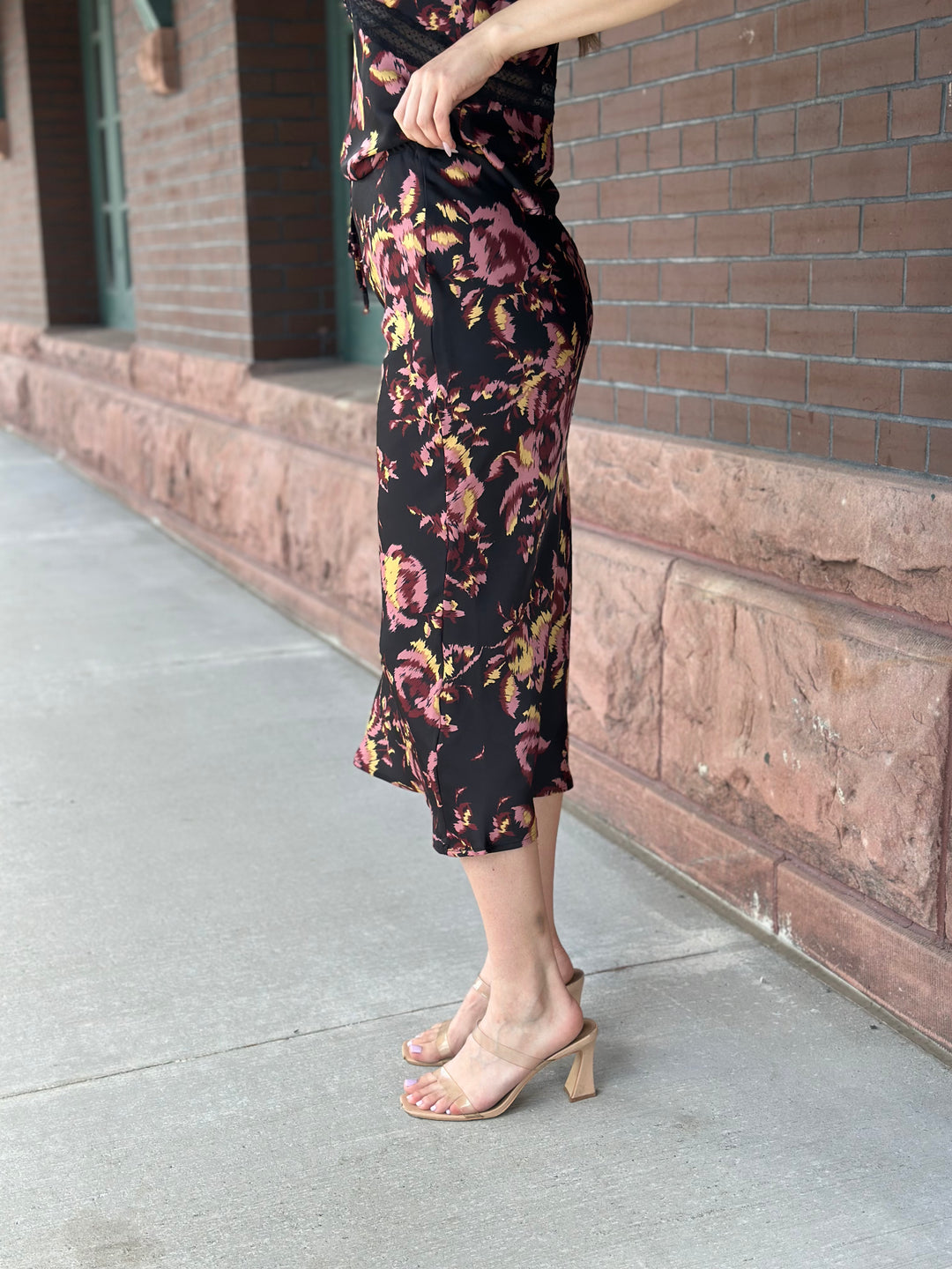 A person wearing a black midi skirt with a pink and yellow floral print, paired with beige block-heel slide sandals, standing on a concrete sidewalk next to a brick building.