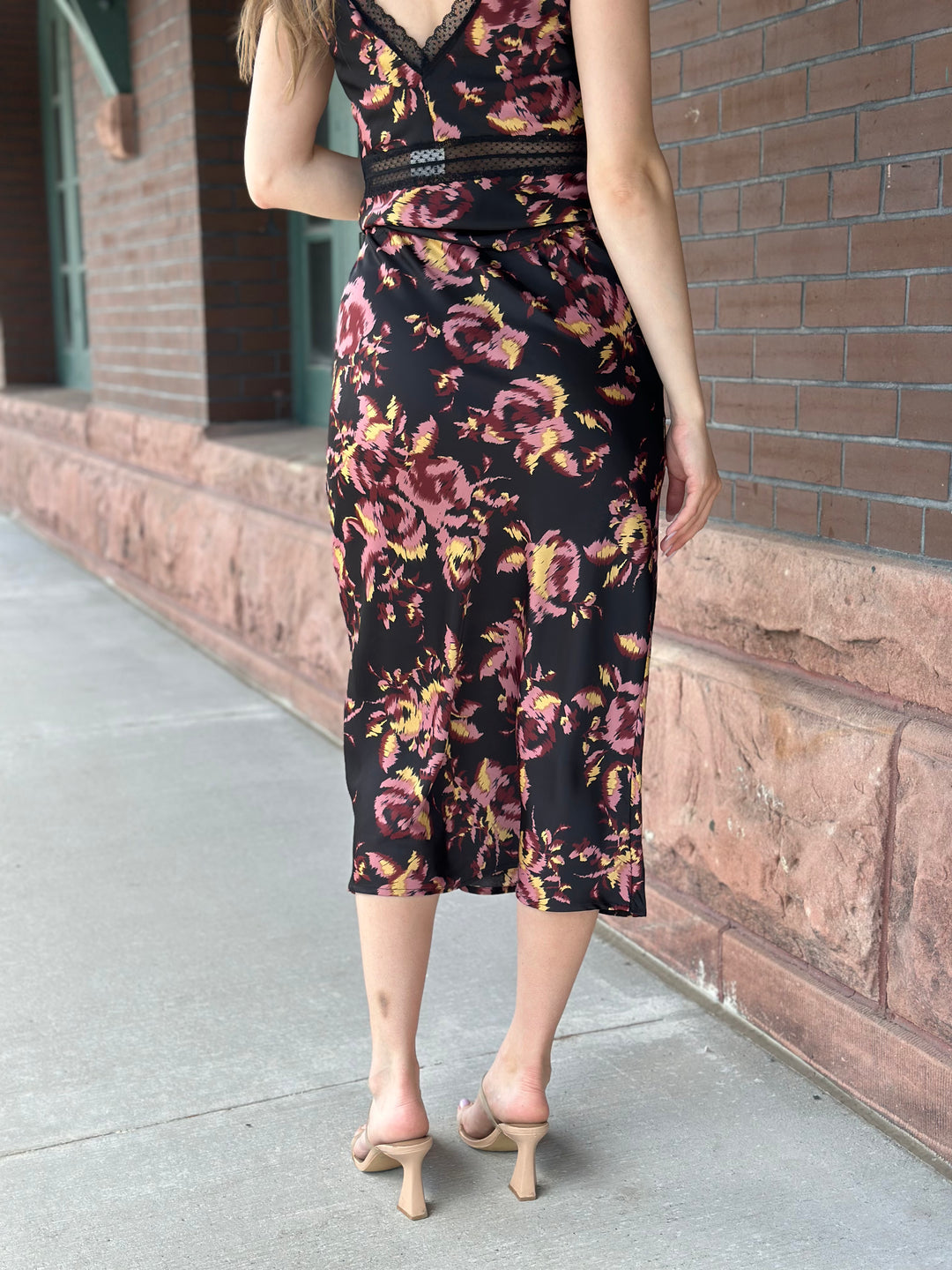 A woman wearing a sleeveless black midi dress with a pink and yellow floral print, shown from the back as she stands on a sidewalk next to a brick building.