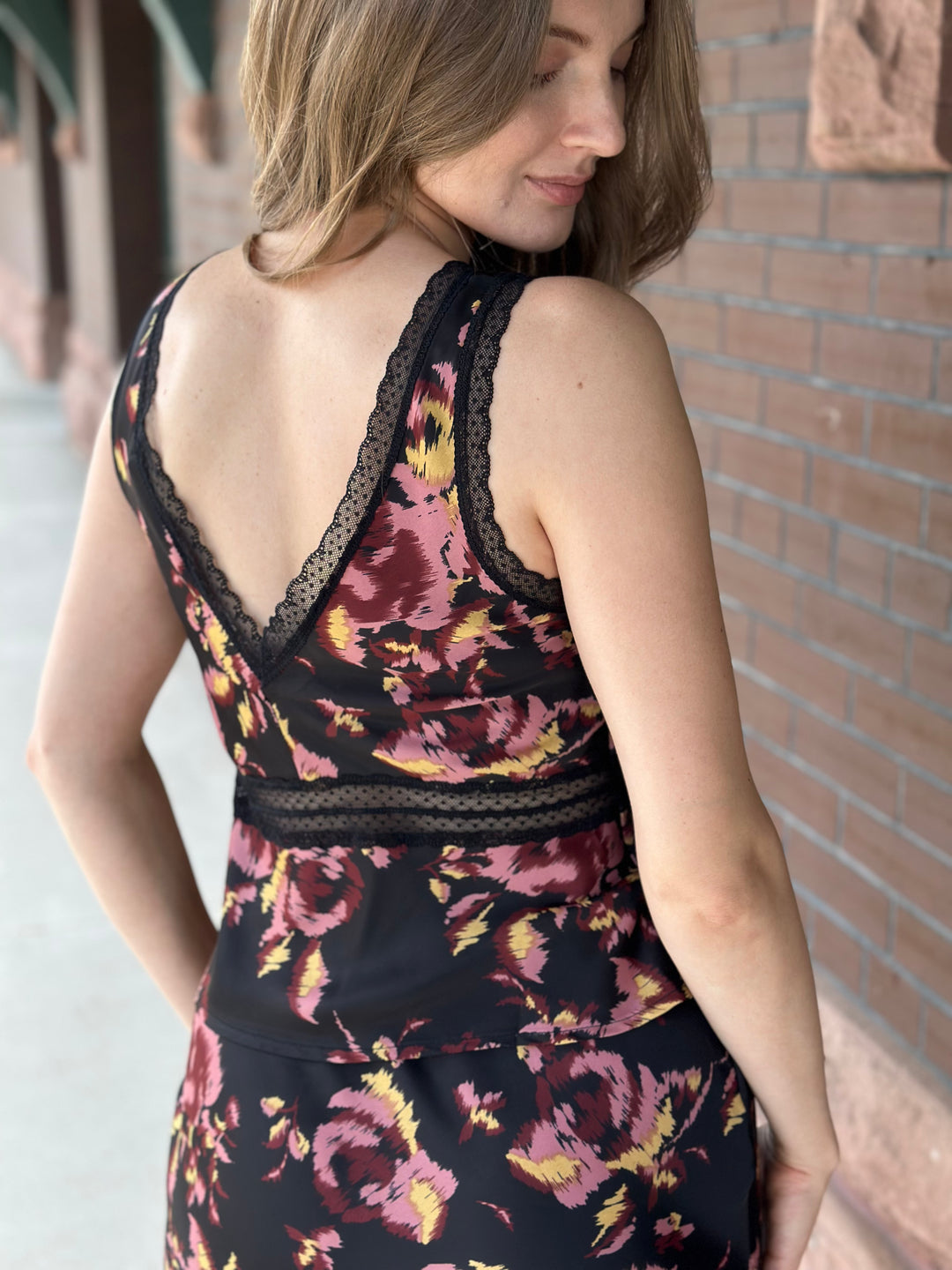 A woman wearing a sleeveless black dress with a pink, red, and yellow floral pattern and black lace trim, shown from the back in an outdoor setting next to a brick wall.