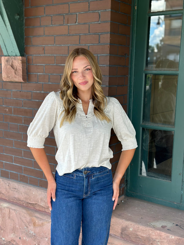 A woman with blonde hair stands outdoors in front of a brick building, wearing a cream-colored short-sleeve button-up blouse and blue jeans.