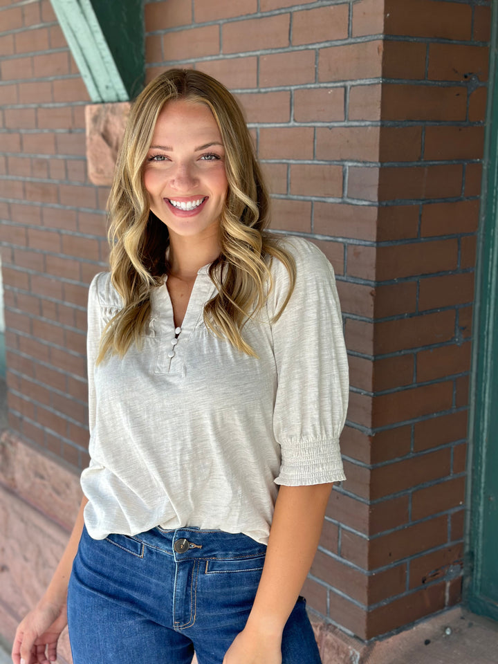 A smiling blonde woman wearing a light beige short-sleeve blouse with puff sleeves and blue jeans, standing in front of a brown brick wall.