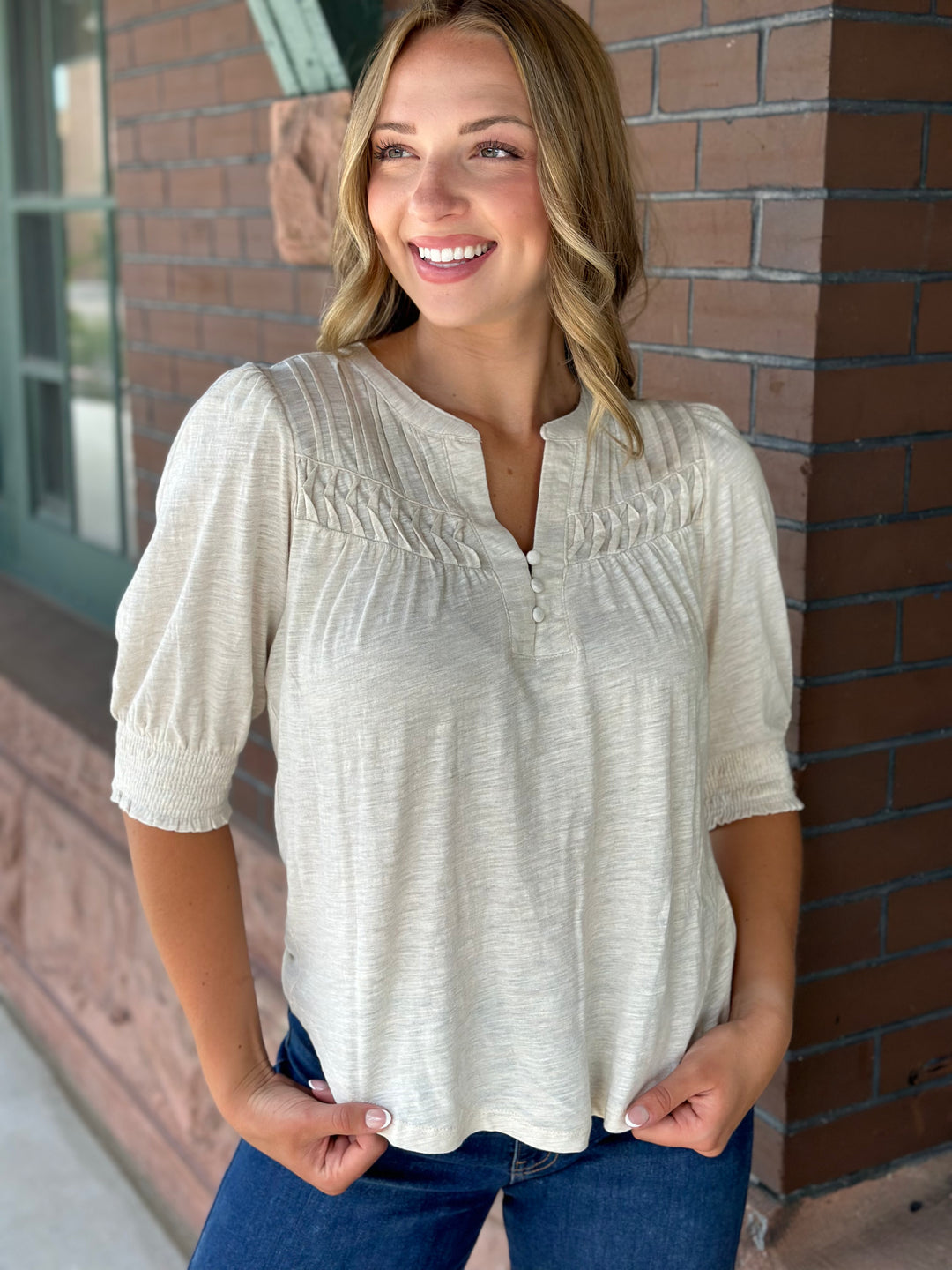 A woman wearing a light beige textured short-sleeve blouse with a smocked chest detail, paired with blue jeans, standing in front of a brick wall.