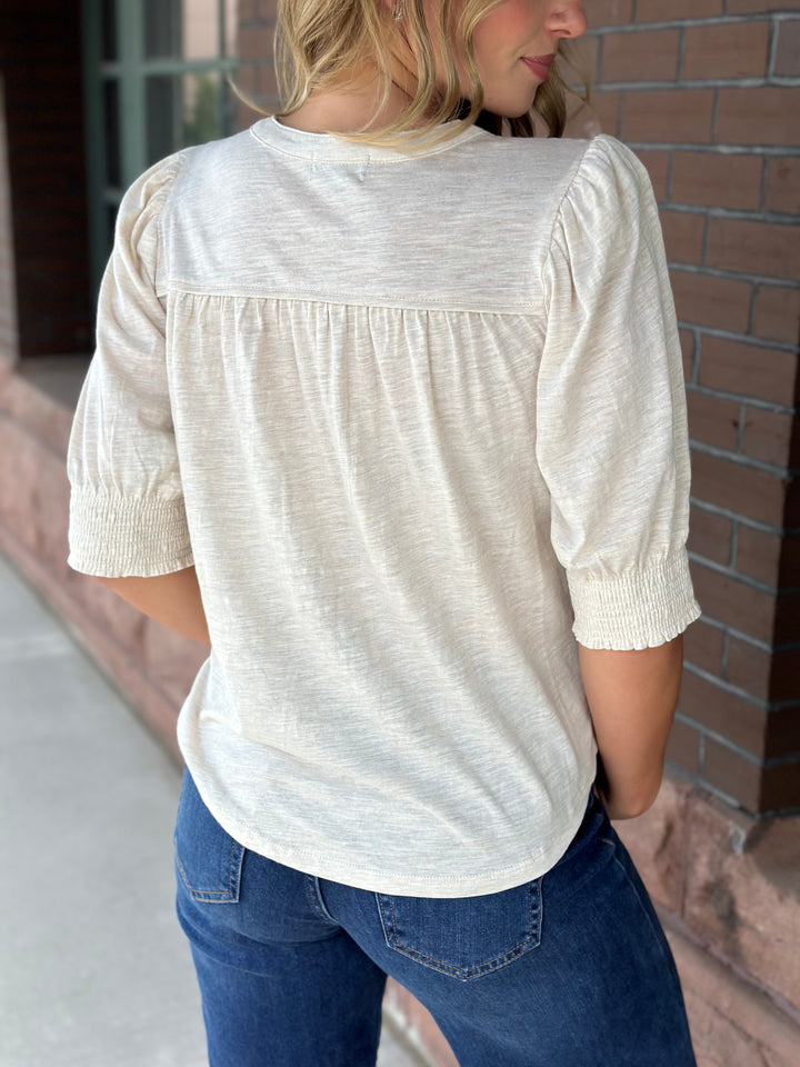 A woman wearing a cream-colored, textured three-quarter sleeve blouse with puff sleeves and a gathered back detail, paired with blue jeans, standing outdoors next to a brick wall.