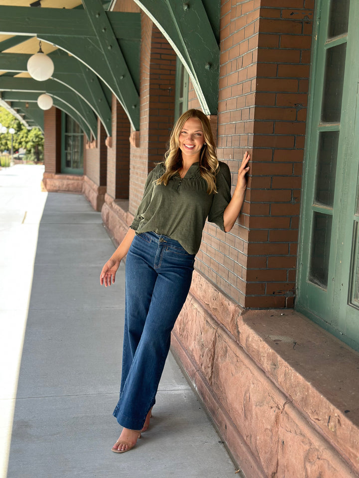 A woman stands barefoot on a covered walkway next to a brick building, wearing an olive green short-sleeve top and blue flare jeans.