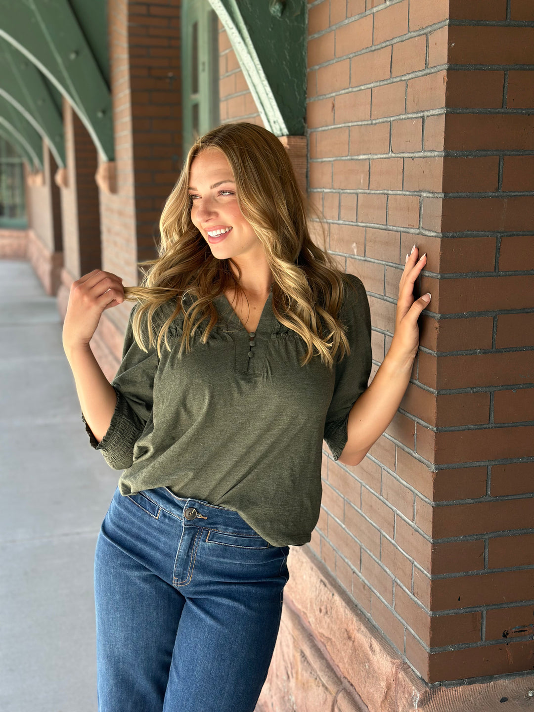 A woman with blonde hair leans against a brown brick column in an outdoor walkway, wearing an olive green short-sleeve top and blue jeans.