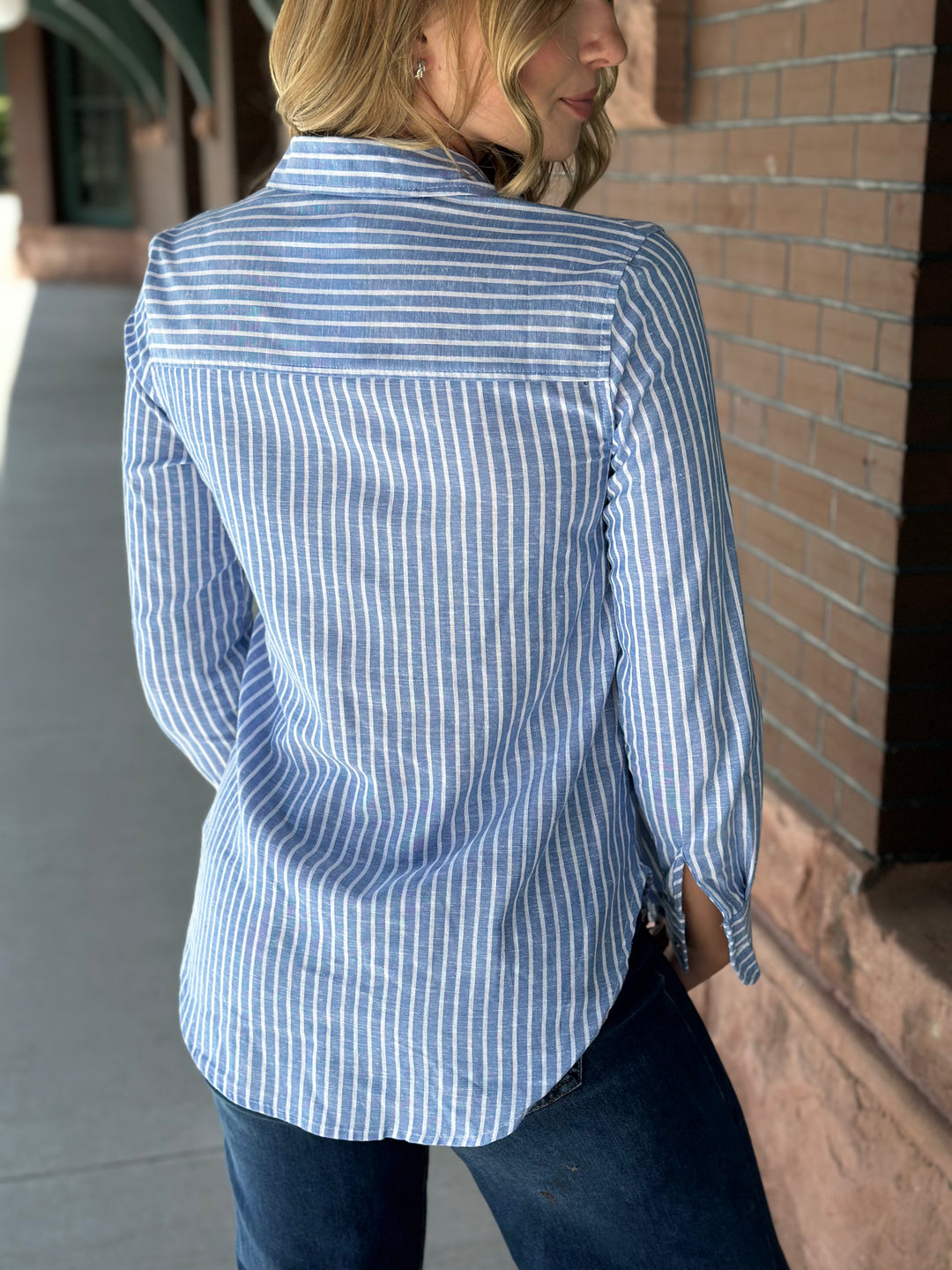 A woman wearing a light blue and white vertically striped button-down shirt, shown from the back as she stands outdoors next to a brick wall.