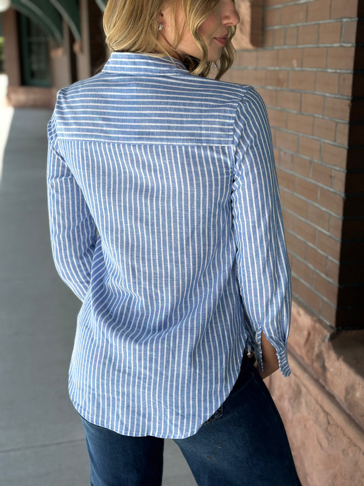 A woman wearing a light blue and white vertically striped button-down shirt, shown from the back as she stands outdoors next to a brick wall.