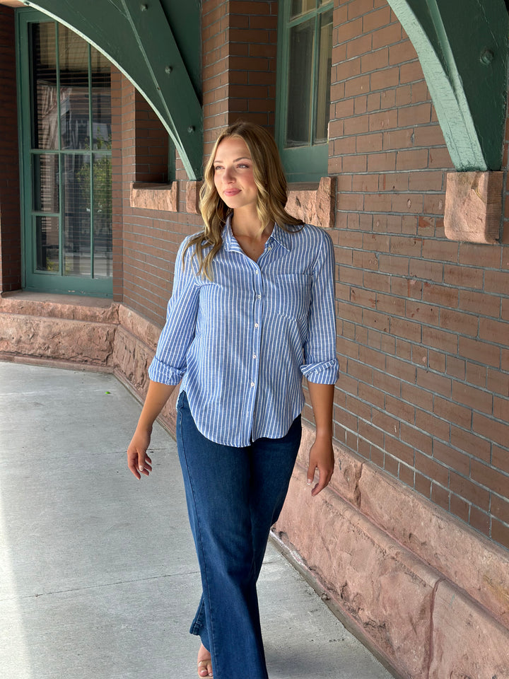A woman wearing a light blue and white vertically striped button-down shirt with rolled-up sleeves and dark blue jeans, standing on a concrete walkway next to a brick building with green metal arches.