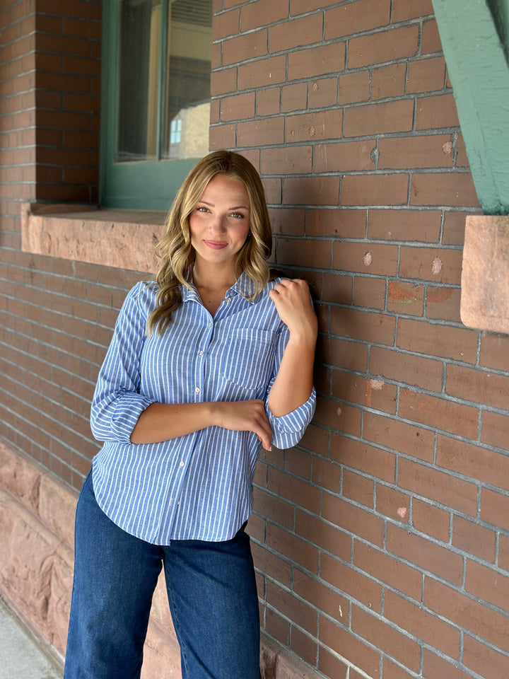 A woman with blonde hair leans against a red brick wall, wearing a light blue and white vertically striped button-down shirt with dark blue jeans.