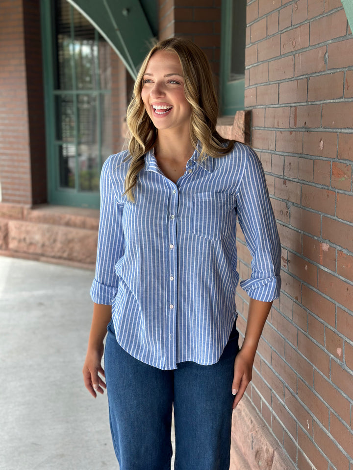 A woman smiling and wearing a blue and white vertically striped button-down shirt with dark blue jeans, standing outdoors against a brick wall.
