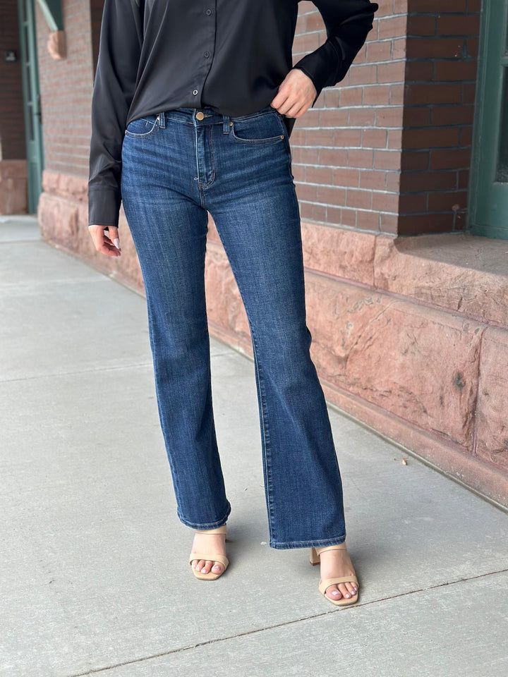 A person wearing dark blue straight-leg or bootcut jeans, a black long-sleeve button-up shirt, and beige heeled sandals, standing on a sidewalk in front of a brick building.