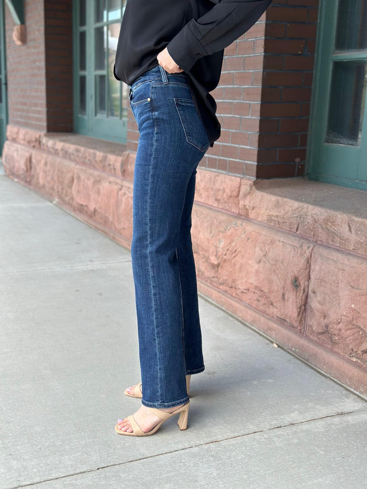 A person wearing dark blue straight-leg or bootcut jeans, a black long-sleeve top, and beige heeled sandals, standing on a sidewalk next to a brick building.