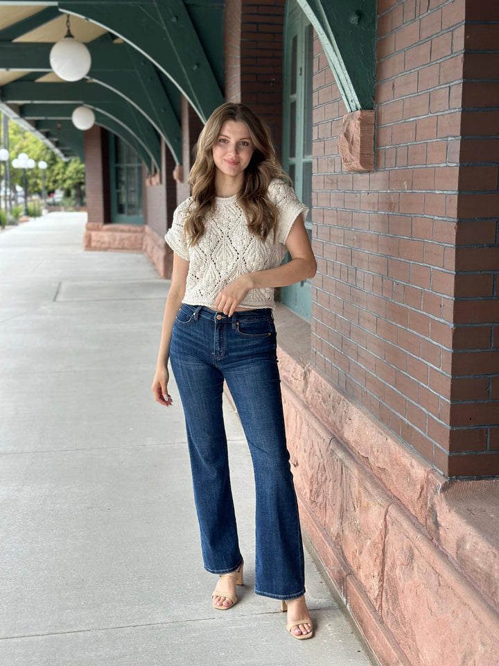 A woman stands on a sidewalk next to a brick building, wearing a cream-colored lace short-sleeve top and dark blue flare jeans.