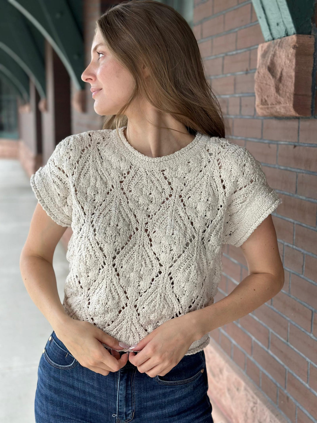 A woman wearing a cream-colored, short-sleeve cable-knit sweater and blue jeans, standing outdoors against a brick wall.