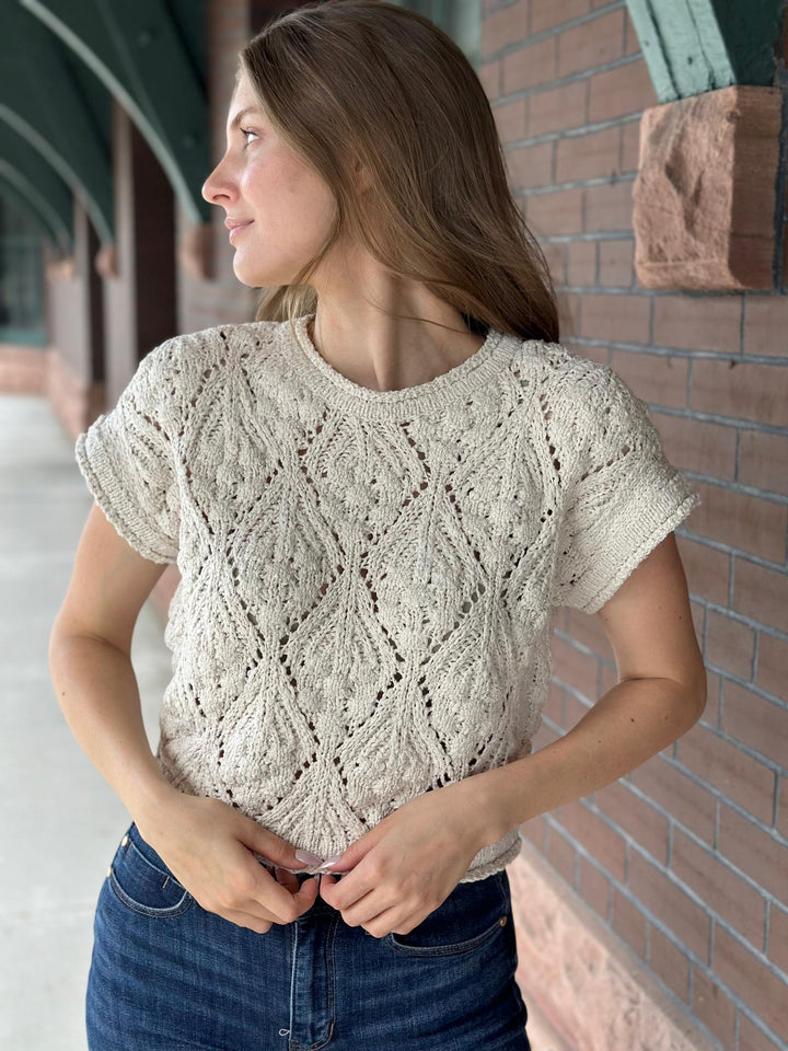 A woman wearing a cream-colored, short-sleeve cable-knit sweater and blue jeans, standing outdoors against a brick wall.