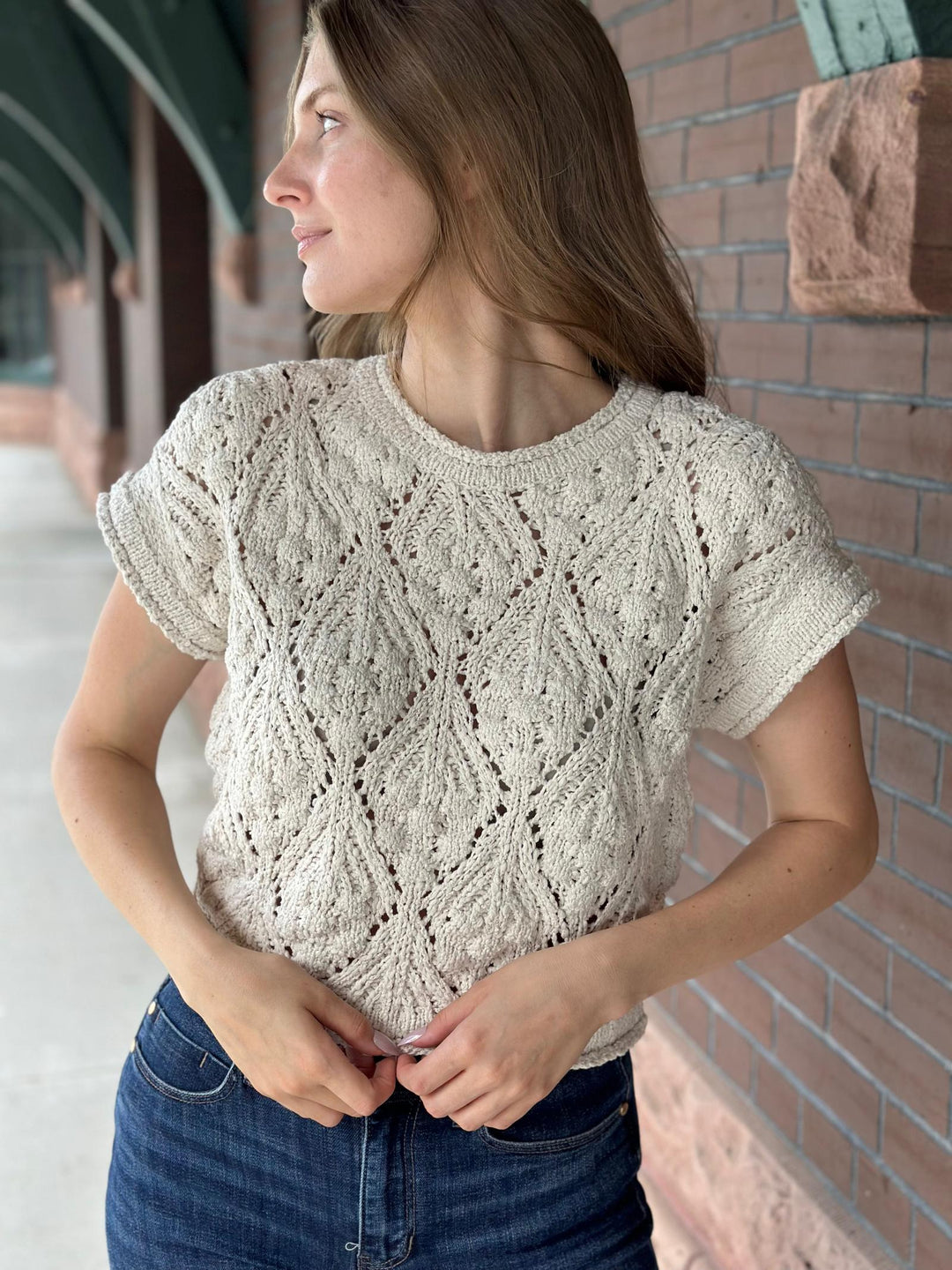 A woman wearing a cream-colored, short-sleeve knit top with an intricate openwork diamond pattern, paired with blue jeans, standing outdoors against a brick wall.