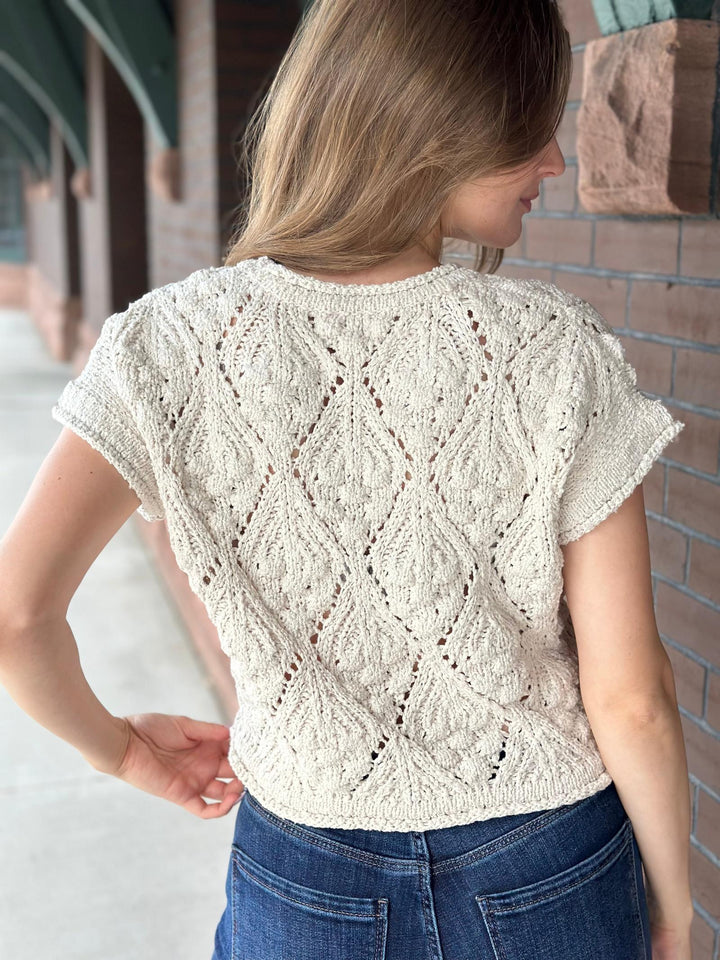 A woman viewed from the back wearing a cream-colored, short-sleeve lace knit top with a textured diamond pattern, paired with blue jeans, standing outdoors against a brick wall.