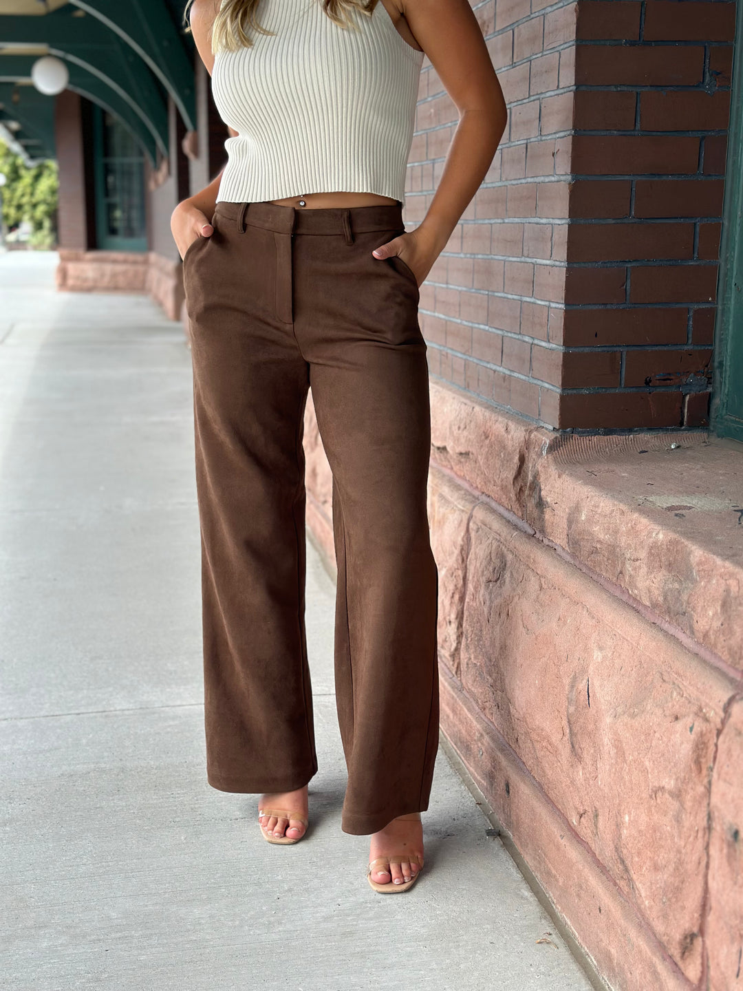 A woman wearing brown wide-leg trousers and a white ribbed tank top stands on a sidewalk beside a brick building.