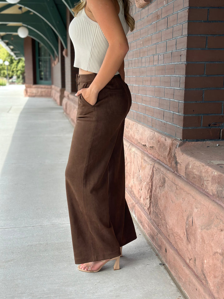 A woman wearing brown wide-leg corduroy pants and a ribbed cream top stands on a sidewalk next to a brick building.