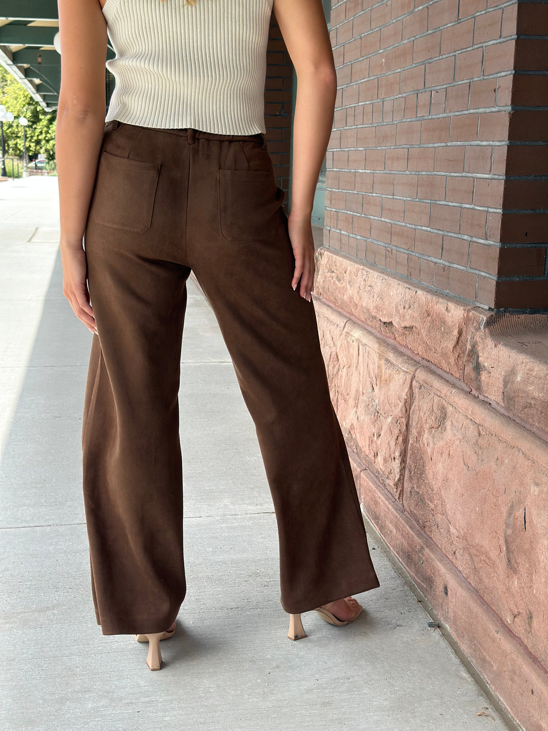 A person wearing high-waisted, wide-leg brown pants and a cream ribbed tank top, standing on a sidewalk next to a brick wall.