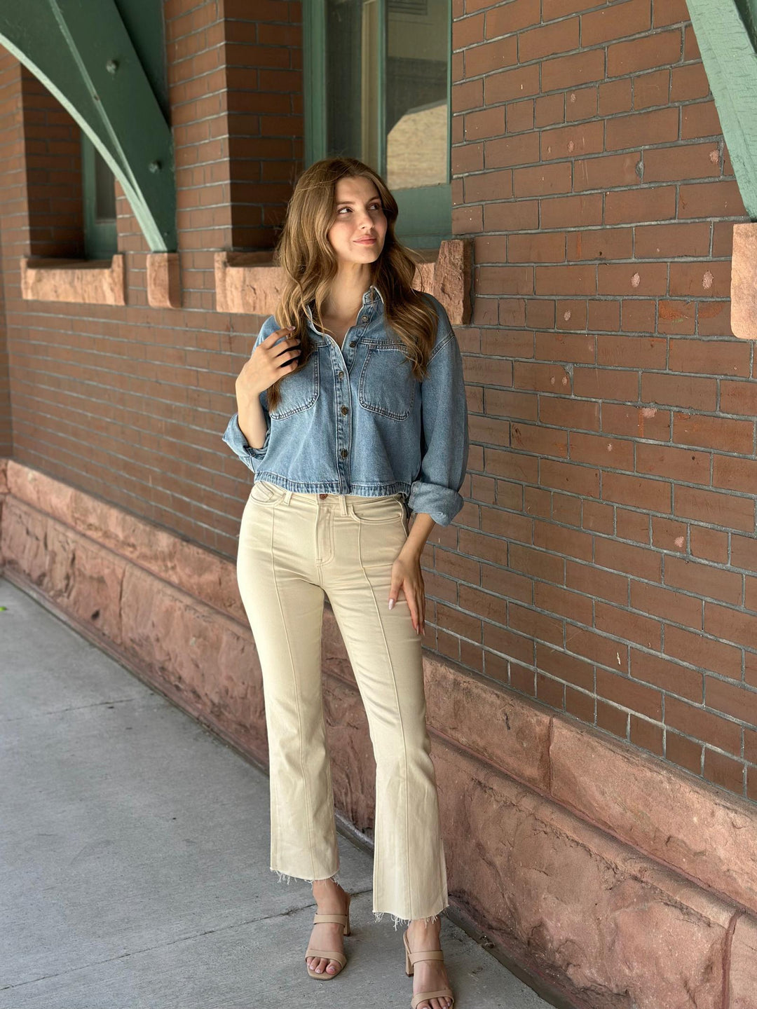 A woman stands outdoors against a red brick building, wearing a light blue cropped denim jacket with rolled sleeves, cream-colored cropped flare jeans, and beige heeled sandals.