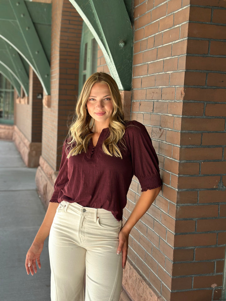 A woman with blonde hair stands in a brick walkway, wearing a burgundy short-sleeve blouse with ruffled trim and cream-colored pants.