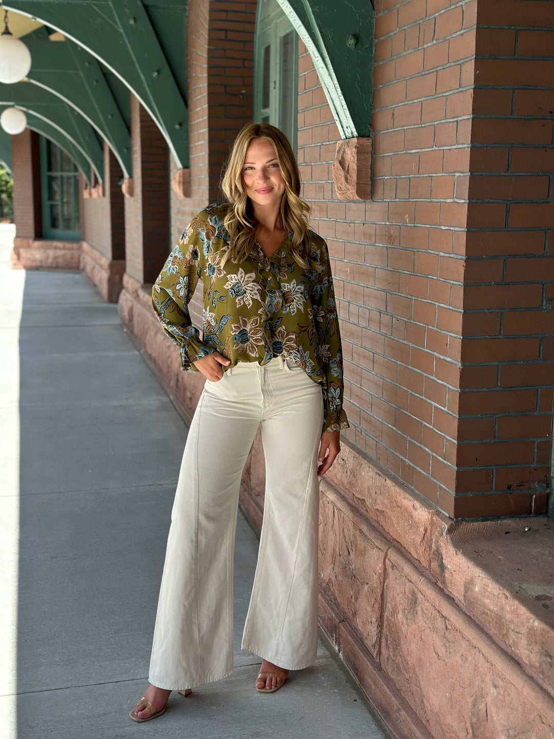A woman stands on a covered walkway wearing a long-sleeve olive green floral blouse and cream wide-leg pants.