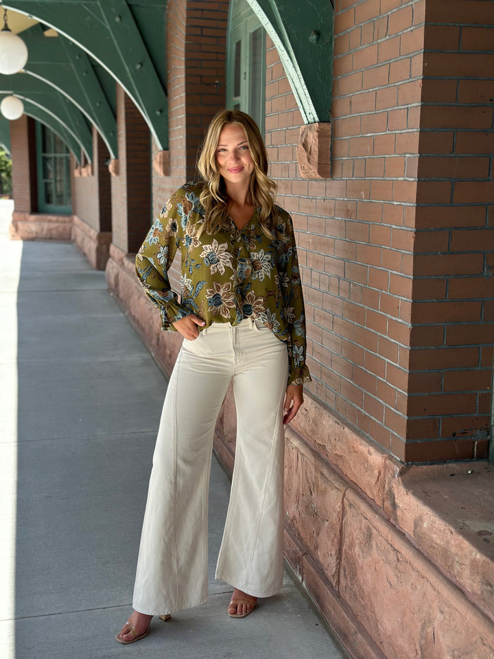 A woman stands on a covered walkway wearing a long-sleeve olive green floral blouse and cream wide-leg pants.