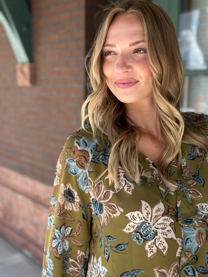 A woman with blonde wavy hair wearing a green floral blouse, standing outdoors in front of a brick wall.