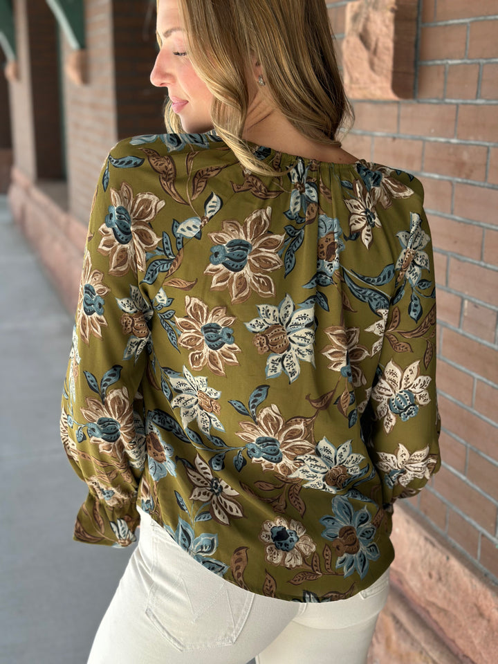 A woman wearing a sage green long-sleeve blouse with a blue, beige, and brown floral pattern, paired with white pants, standing outdoors against a brick wall.