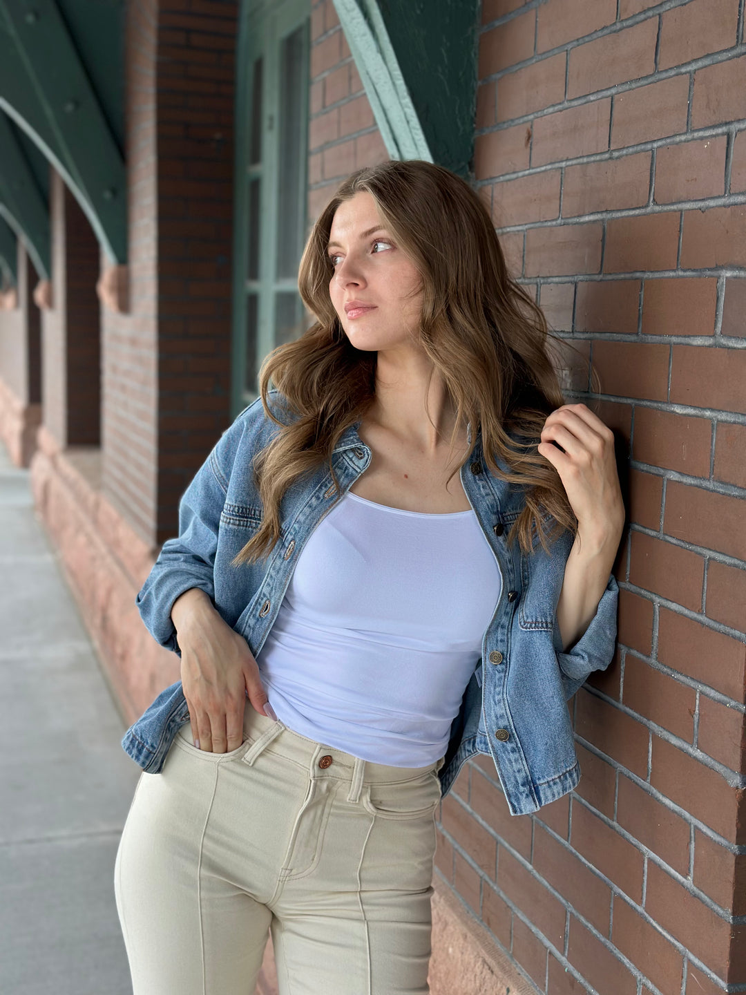 A woman with long brown hair leans against a red brick wall, wearing a light blue denim jacket over a white ribbed tank top and cream-colored jeans.